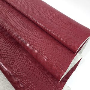 Peut inclure: Cuir synthétique bordeaux avec un motif peau de serpent. Le cuir est plié en trois, montrant la texture du matériau.