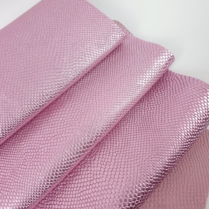 Può includere: Primo piano di diversi fogli di finta pelle rosa con un motivo strutturato a scaglie. Il materiale ha una lucentezza metallica, che riflette la luce. I fogli sono piegati, rivelando il lato inferiore del materiale. Lo sfondo è bianco.