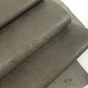 May include: Close-up of several sheets of embossed leather in a muted olive green color. The leather has a detailed floral pattern. The sheets are folded and layered, revealing the texture and color variations of the material.