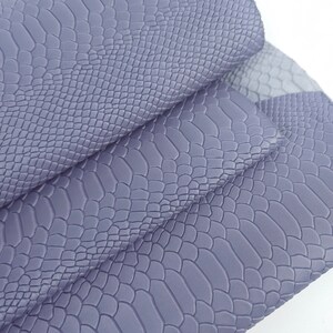 Pode incluir: Um close-up de um tecido de imitação de pele de cobra roxo. O tecido tem um padrão texturizado e em relevo que se assemelha a escamas de cobra.