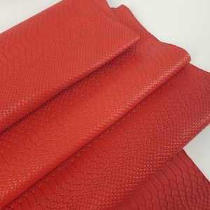 May include: A folded piece of bright red leather featuring a textured snakeskin pattern across the surface. The material is layered, showing the embossed scale design clearly on the top and bottom sections of the vibrant red hide.