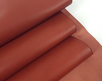 May include: Close-up of several sheets of reddish-brown leather. The leather is folded over, revealing a smooth, finished surface. The edges of the leather are visible, showing the thickness and texture. The background is white.