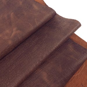 Könnte beinhalten: Nahaufnahme von mehreren Stücken braunem Leder. Das Leder hat eine strukturierte Oberfläche mit Farb- und Musterunterschieden. Die Farbtöne reichen von einem tiefen, satten Braun bis zu einem helleren, rötlichen Braun. Das Leder scheint gefaltet und geschichtet zu sein.