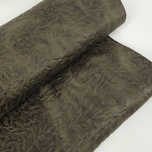 May include: Close-up of a textured, olive-green leather material. The leather has a crinkled appearance with a leaf-like pattern embossed across the surface. The material is folded over, revealing more of the textured surface.