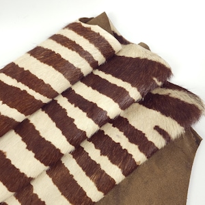May include: Close-up of zebra-striped hide with alternating bands of brown and cream. The hide is layered over a piece of brown suede-like material. The texture of the zebra stripes is visible, suggesting a natural animal skin.