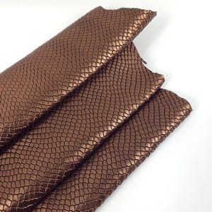 Op de afbeelding: Close-up van verschillende vellen bruin imitatieleer met een textuur, schubachtig patroon. Het materiaal is gevouwen, waardoor de onderkant en randen zichtbaar zijn. De belichting benadrukt de metallic glans van het materiaal.