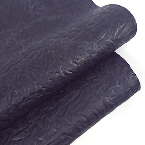 May include: A close-up of a piece of dark navy blue leather with a textured, wrinkled surface. The leather is folded over, revealing the underside. The material has a matte finish and appears to be high-quality.