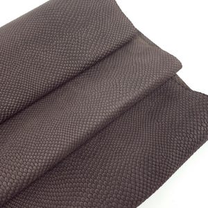 Puede incluir: Primer plano de un trozo de cuero marrón doblado, que muestra un patrón texturizado en forma de escamas. El cuero tiene un acabado mate y parece suave. La imagen está bien iluminada, destacando los detalles del material. El texto "Leatherforcraft" es visible.