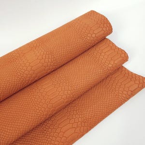 Puede incluir: Primer plano de una pieza de cuero naranja doblada con un patrón en relieve de cocodrilo. El cuero es suave y flexible, y el patrón es realista.