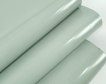 May include: Close-up of three rolls of light gray, glossy vinyl material. The material has a smooth, reflective surface and appears to be rolled up. The rolls are stacked on top of each other, with the top roll partially unrolled.