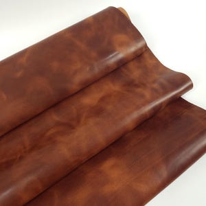 Puede incluir: Una pieza grande y doblada de cuero de color marrón medio con un acabado suave y mate. La superficie muestra variaciones naturales de color, que van desde el caoba profundo hasta tonos bronceados más claros, creando un aspecto jaspeado.