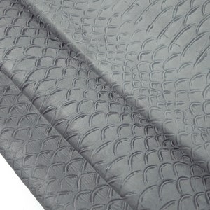 Peut inclure: Gros plan sur un tissu gris avec un motif texturé en relief ressemblant à des écailles. Le tissu est plié en deux, montrant le motif des deux côtés.