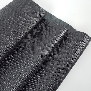 Könnte beinhalten: Nahaufnahme von schwarzem Lederstoff mit einem strukturierten, schuppenartigen Muster. Das Material ist gefaltet, wodurch mehrere Schichten und ein Blick auf die Unterseite sichtbar werden. Die dunkle Farbe und die detaillierte Textur deuten auf ein hochwertiges Material hin.