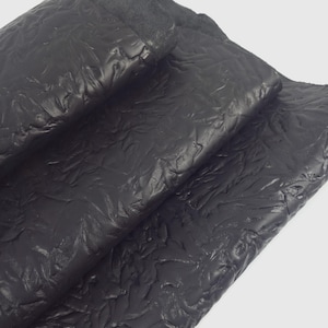 May include: Close-up of black leather with a textured, embossed pattern. The leather is folded over itself, revealing the underside. The surface has a raised, organic design, creating a tactile and visual appeal. The material appears to be high-quality.