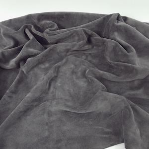 May include: A close-up of a draped, dark grey fabric. The material appears soft, possibly velvet or suede, with visible folds and shadows. The fabric is spread out, creating a textured surface.