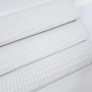 May include: White faux leather fabric with a crocodile embossed pattern. The fabric is folded in thirds.