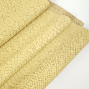 Könnte beinhalten: Nahaufnahme von mehreren Bahnen hellgelben Kunstleders mit einem Reptilienschuppenmuster. Die Bahnen sind gefaltet und geschichtet, wodurch die Textur- und Farbvariationen sichtbar werden. Das Material scheint weich und flexibel zu sein, geeignet für Bastelarbeiten oder Polsterungen.