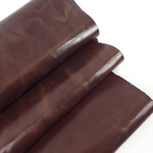 Puede incluir: Primer plano de varias hojas de cuero marrón oscuro. El cuero tiene una superficie lisa y brillante con sutiles variaciones de color y textura. Los bordes del cuero son visibles, mostrando el grosor y la calidad del material.