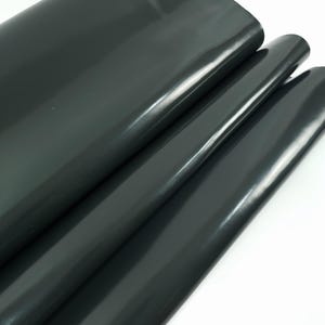 May include: Dark gray, glossy vinyl fabric folded over itself. The material has a smooth, reflective surface, with subtle highlights and shadows. The fabric appears to be thick and durable, suitable for various crafting projects.