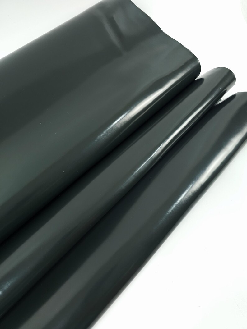 May include: Dark gray, glossy vinyl fabric folded over itself. The material has a smooth, reflective surface, with subtle highlights and shadows. The fabric appears to be thick and durable, suitable for various crafting projects.