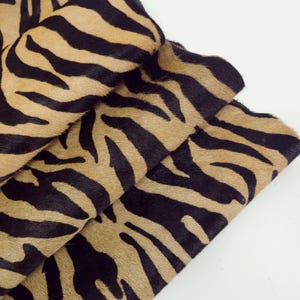 Op de afbeelding: Close-up van gevouwen stof met zebra-strepen. De stof heeft een bruinachtige achtergrond met dikke zwarte strepen. Het materiaal lijkt zacht en getextureerd, met een natuurlijk dierenprintpatroon. De stof is over zichzelf gevouwen.