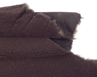 May include: Close-up of a section of brown shearling fabric. The fabric has a dense, textured surface on one side and a smooth, dark brown leather-like backing. The edges are uneven, showing the natural cut of the material.