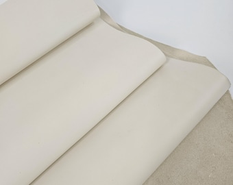 May include: Close-up of cream-colored leather sheets, rolled and stacked, with a rough, textured surface visible on the bottom right. The smooth, folded leather contrasts with the raw, unfinished edge, suggesting a material for crafting.