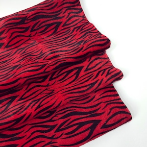 May include: A piece of red and black zebra-striped cowhide leather. The leather has a soft, textured surface with a bold pattern of black stripes against a vibrant red background. The material is folded over, revealing the texture and color.