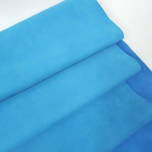 Pode incluir: Uma pilha de três camadas de tecido azul em tons variados. A camada superior é um turquesa brilhante, seguida por um azul médio e um azul profundo na parte inferior. O tecido parece macio e liso.
