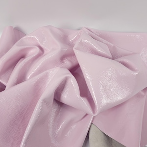 May include: A close-up of a sheet of pink patent leather. The leather is folded and draped, showing its shiny, textured surface. The underside of the leather is a light beige colour.