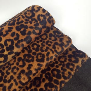 May include: Rolled leopard print fabric with a brown and black spotted pattern. The fabric has a soft, textured appearance and is partially rolled, revealing the underside of the material, which is a dark grey colour.