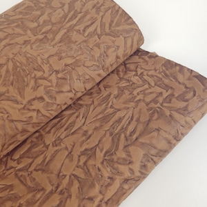 May include: Two sheets of brown textured fabric with a leaf-like pattern. The fabric is folded over, revealing the underside. The material has a rough, crinkled appearance, and the color is a warm, earthy brown.