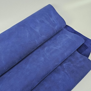 以下が含まれることがあります： 濃い青色の革のシートのクローズアップ。革は柔らかく、スエードのような質感で、色とトーンに微妙な変化があります。折り目と折り目は、素材に深さと奥行きを追加します。