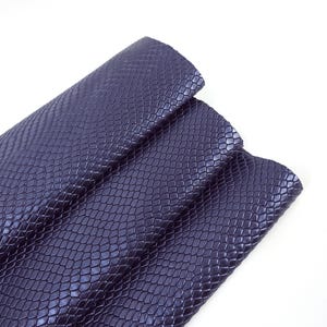 Può includere: Primo piano di un materiale viola scuro, strutturato con un motivo a pelle di serpente. Il materiale è piegato, rivelando la dettagliata texture a scaglie. Lo sfondo è bianco, evidenziando il colore ricco e il motivo del tessuto.