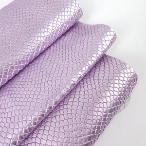 Op de afbeelding: Drie rollen lavendelkleurig imitatieleer. Het materiaal heeft een textuur, reptielachtig patroon. De rollen zijn op elkaar gestapeld, waarbij de bovenste rol gedeeltelijk is uitgerold en de onderkant zichtbaar is.