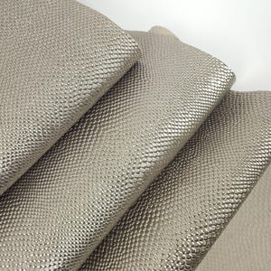 May include: Close-up of several sheets of metallic silver-grey faux leather. The material has a textured, scaled pattern, and the folds of the fabric are visible. The lighting creates highlights and shadows, emphasising the texture and sheen of the material.
