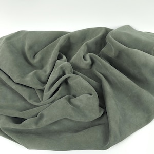 May include: A draped piece of olive green fabric against a white background. The fabric is soft and appears to be suede or a similar material. The folds and shadows create a sense of depth and texture. The fabric is likely used for clothing or home decor.