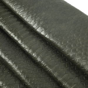 May include: Close-up of a dark green leather fabric with a textured, scaly pattern. The fabric is folded in several layers, revealing the texture.