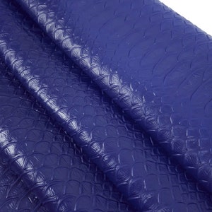 Op de afbeelding: Een close-up van een blauwe kunstlederen stof met een textuur van schubben. De stof is gevouwen en gedrapeerd, waardoor de textuur en diepte van het materiaal zichtbaar worden.