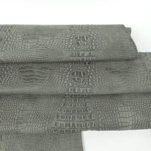 May include: Close-up of several sheets of gray crocodile-textured leather. The leather has a detailed, scaled pattern and a matte finish. The sheets are stacked, revealing the texture and color variations.