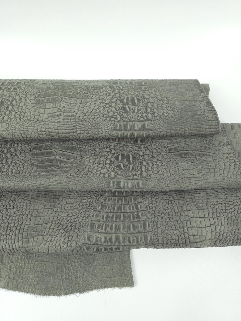 May include: Close-up of several sheets of gray crocodile-textured leather. The leather has a detailed, scaled pattern and a matte finish. The sheets are stacked, revealing the texture and color variations.