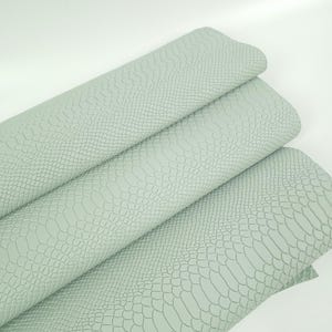 Puede incluir: Un rollo de tela de piel sintética de color verde salvia claro con un patrón texturizado de piel de serpiente. El material se muestra en tres pliegues distintos, exhibiendo el diseño de escamas en relieve en toda la superficie de la hoja.