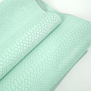 Può includere: Primo piano di fogli di ecopelle verde menta con un motivo a squame di rettile. Il materiale è piegato, rivelando la consistenza e le variazioni di colore. I fogli sono probabilmente utilizzati per progetti di artigianato o tappezzeria.