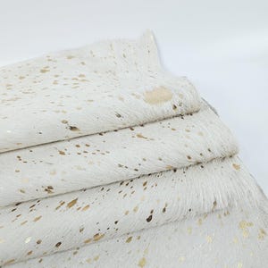 Peut inclure: Gros plan sur un tapis en peau de vache blanche avec des éclaboussures dorées métalliques. Le tapis est plié sur lui-même, montrant la texture et le motif. La peau de vache a une texture douce et duveteuse.