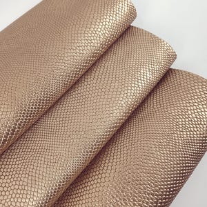 May include: Close-up of three sheets of metallic gold faux leather. The material has a textured, scale-like pattern. The sheets are folded over each other, revealing the shiny surface and the detailed texture.