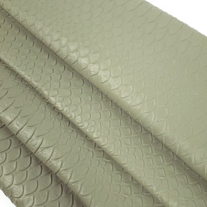 Può includere: Primo piano di un tessuto effetto pelle di serpente grigio chiaro con un motivo testurizzato e squamoso. Il tessuto è piegato in più strati, mostrando la trama.