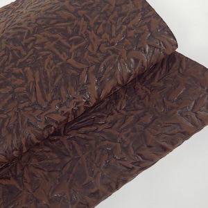 May include: Close-up of a folded piece of brown leather with a textured, wrinkled surface. The leather has a dark, rich color and appears to be pliable. The texture creates a pattern of raised and recessed areas.