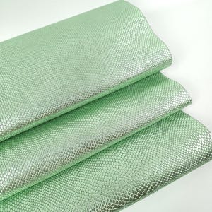 May include: Three sheets of mint green faux leather with a textured, scale-like pattern. The material has a subtle sheen, reflecting light. The sheets are layered, showcasing the texture and color variations.