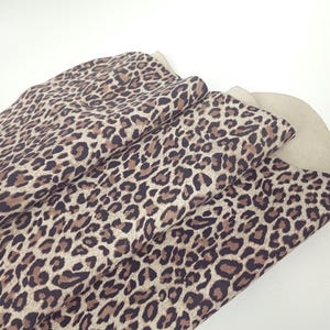 Könnte beinhalten: Ein gefaltetes Stück Stoff mit einem sich wiederholenden Leopardenmuster in den Farben Dunkelbraun, Hellbraun und Creme. Das Textil zeigt ein klassisches Tierfellmotiv mit unregelmäßigen dunklen Flecken auf einem hellen Hintergrund.