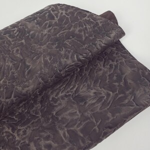 May include: A folded piece of fabric with a textured, abstract pattern in shades of brown and gray. The fabric appears to be soft and pliable, with a slightly shiny surface. The pattern resembles a network of veins or cracks.